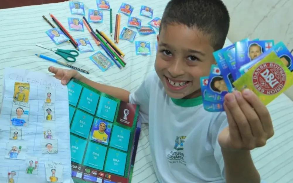 O menino João Gabriel, de 8 anos, com o álbum da Panini e o que desenhou: "Gosto dos dois" - Foto: Fábio Lima/O Popular